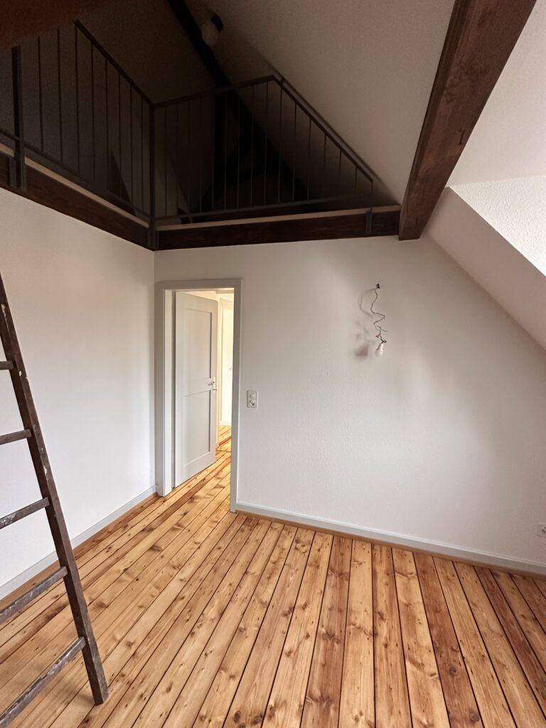 Helles Zimmer im Dachgeschoss mit frisch geschliffenem und geöltem Holzparkett sowie einer Galerie mit Metallgeländer.